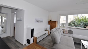 A modern living room with a grey sofa, wooden coffee table, and a white entertainment unit. There is a window with a view of greenery outside.