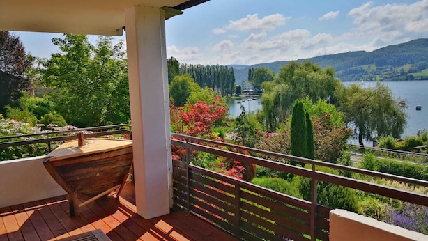 A wooden deck with a bench and a table, overlooking a lake with boats, surrounded by greenery and hills.