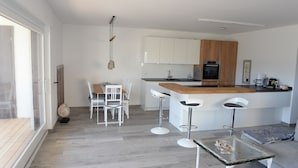 A modern kitchen with white cabinets, a wooden countertop, and a built-in oven. There are three white bar stools and a small table with a glass top.