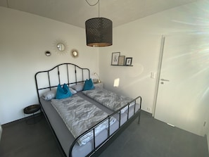 Ein Einzelbett mit Metallgestell, grauer Bettwäsche und blauen Kissen. An der Wand hängt ein Spiegel, darüber eine Pendelleuchte und darunter eine kleine Ablagefläche mit gerahmten Bildern.
