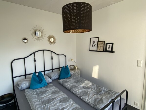 Una cama individual con estructura metálica, ropa de cama gris estampada y dos almohadas azules. Se observa una estantería montada en la pared con cuadros enmarcados y un espejo decorativo.
