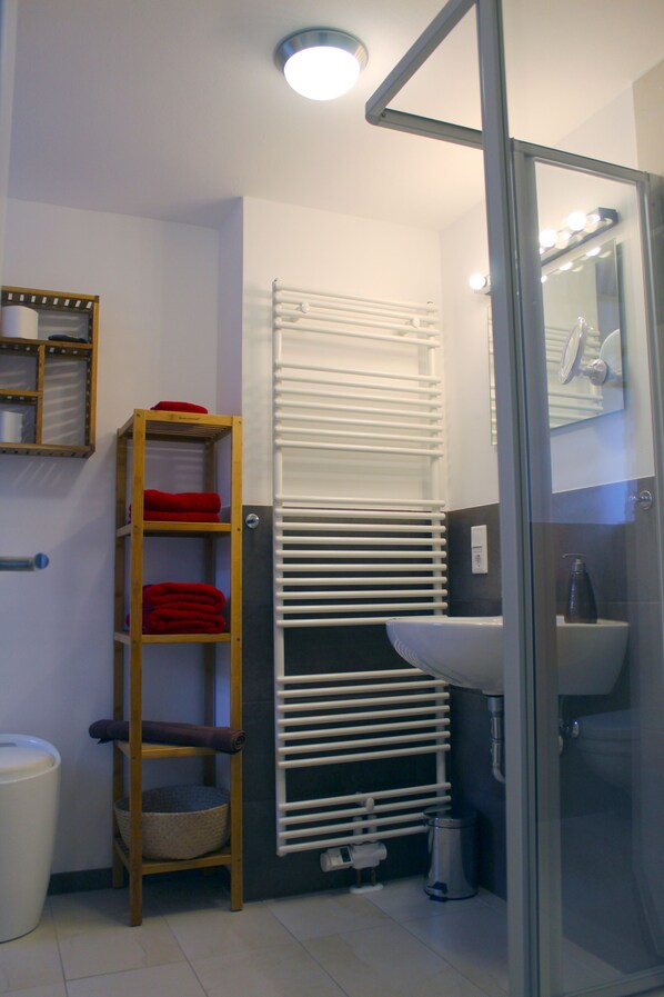 Un baño con un estante de madera con toallas, un radiador blanco, una cabina de ducha de cristal y un lavabo con un espejo encima.