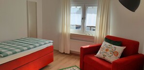 Une pièce avec un lit rouge, un fauteuil rouge orné d’un coussin à motifs floraux, une fenêtre avec des rideaux et un radiateur.
