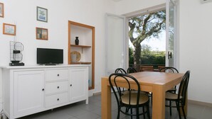 Een eethoek met een houten tafel en stoelen, een wit kastje met een televisie, en een deur naar een balkon met uitzicht op een boom.