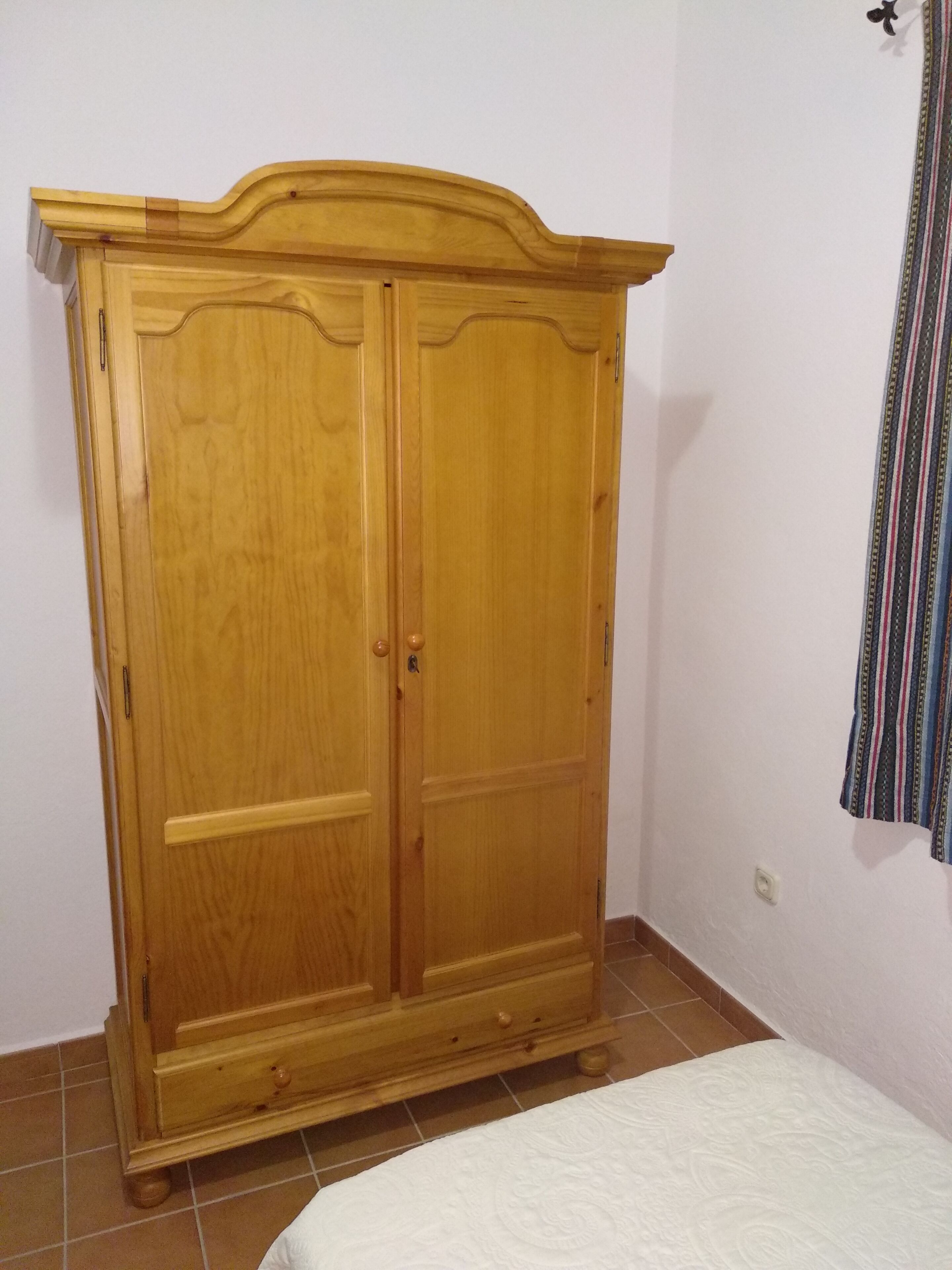 Un armario de madera con dos puertas y un cajón pequeño, ubicado en una habitación con cama y cortina.