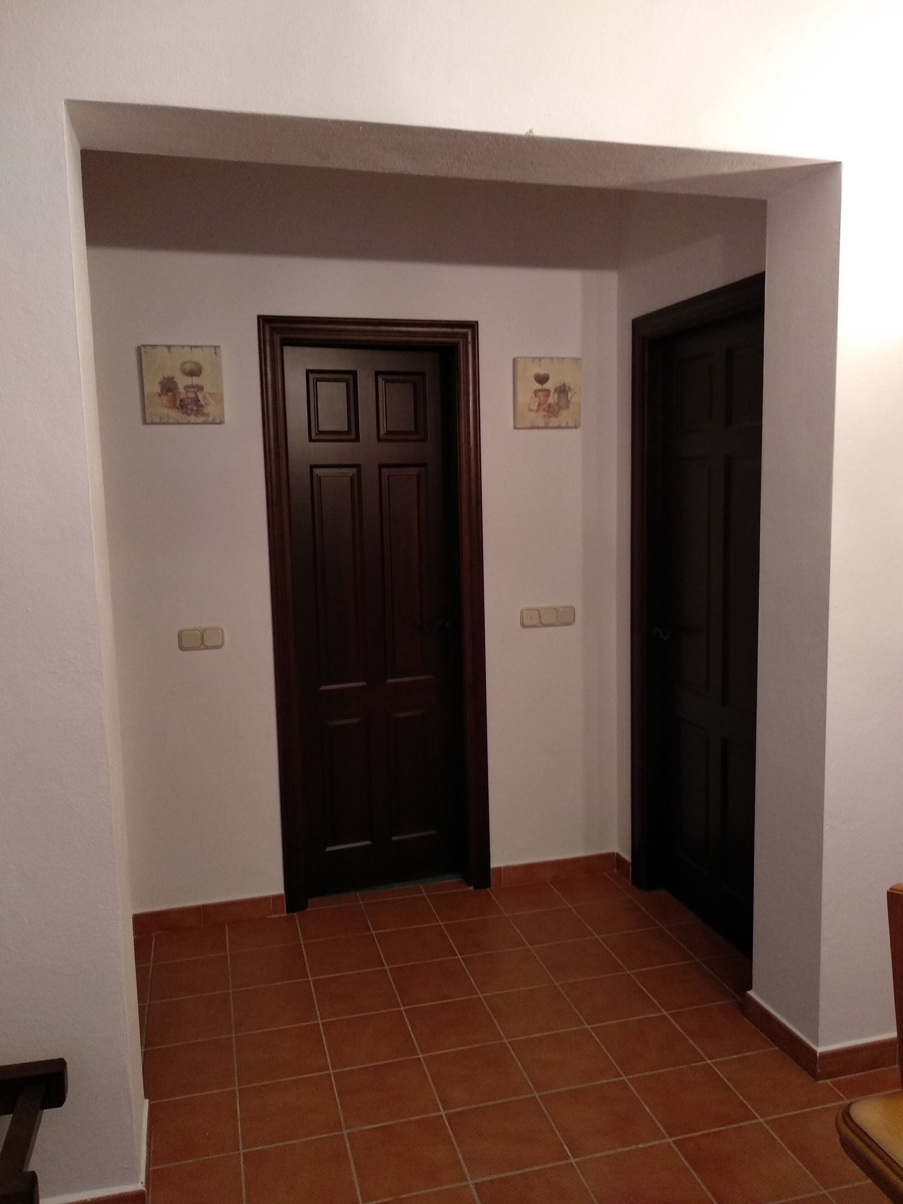 Un pasillo con dos puertas de madera oscura, paredes blancas y suelo de baldosas.