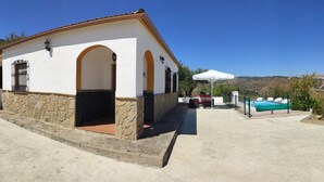 Un bâtiment avec une base en pierre, des murs blancs et un toit recouvert de tuiles rouges. On y trouve un espace piscine avec des chaises longues et un parasol blanc.