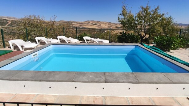 Une piscine rectangulaire à l’eau d’un bleu limpide, entourée de chaises longues blanches et d’une clôture verte.
