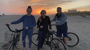Drei Personen stehen an einem sandigen Strand mit Fahrrädern, ein Sonnenuntergang ist im Hintergrund zu sehen.