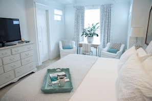 A bedroom with a bed, a dresser, a television, a small table with a plant, two chairs, and a window with curtains.