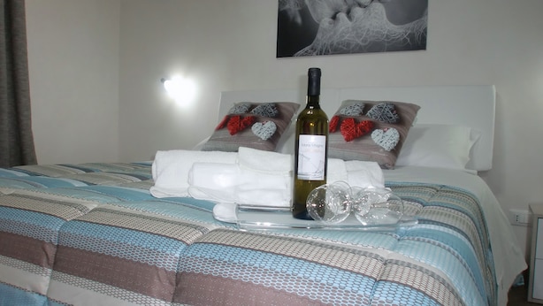 Una cama bien hecha con una colcha estampada, una botella de vino y almohadones con forma de corazón.