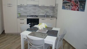 Una cocina compacta con armarios blancos, estufa a gas y un lavamanos. Hay una mesa de comedor con sillas y un florero con flores.