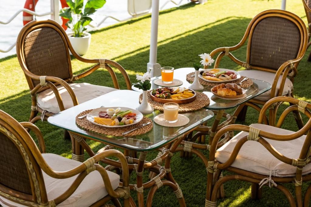 A glass-top table with wicker chairs, surrounded by plates of food and drinks, set on a grassy area.