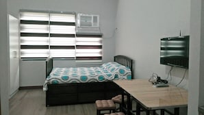 Una habitación pequeña con una cama, un escritorio, una silla, un televisor y un equipo de aire acondicionado.
