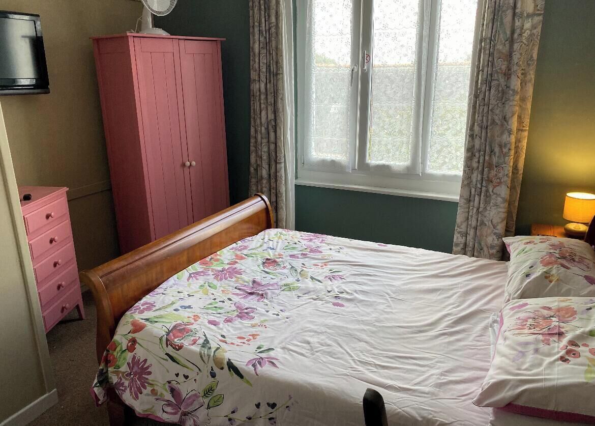 Quarto com cama, guarda-roupa rosa, penteadeira de madeira, ventilador e janela com cortinas.