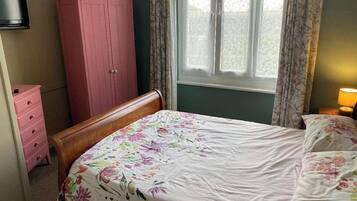 Quarto com cama, guarda-roupa rosa, penteadeira de madeira, ventilador e janela com cortinas.