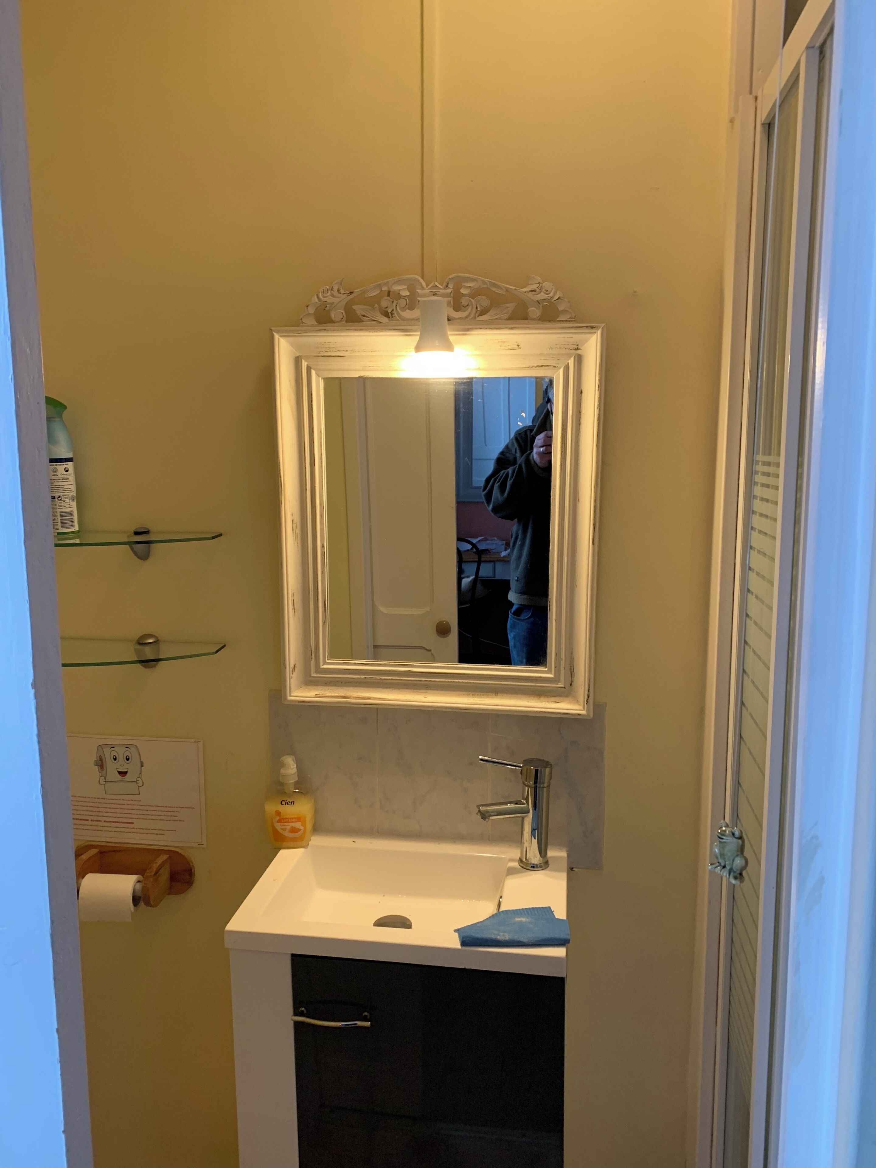 Une salle de bain avec un lavabo blanc, un miroir surmonté d’une lampe et une porte dotée d’un panneau en verre.