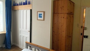 A bedroom with a wooden wardrobe, a bed with a white headboard, a window with blue curtains, and a wall-mounted fan. - Webb's of Glomel (Glomel)