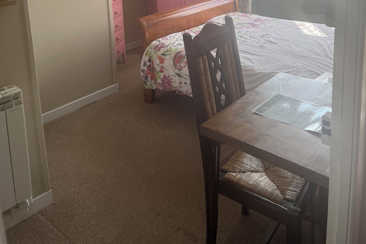A bedroom with a bed, a wooden table, a chair, a pink wardrobe, a ceiling fan, a window with curtains, and a framed picture on the wall.