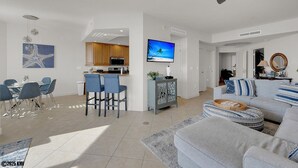 A modern living room with a grey sofa, a dining area with blue chairs, a kitchen with wooden cabinets, and a TV on the wall. - Island Vacation Condo w/ Breathtaking Views At Waterside! First-Class Amenities! (Fort Myers Beach)