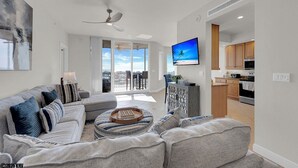A modern living room with a grey sofa, a flat-screen TV, and a balcony with a view. - Island Vacation Condo w/ Breathtaking Views At Waterside! First-Class Amenities! (Fort Myers Beach)
