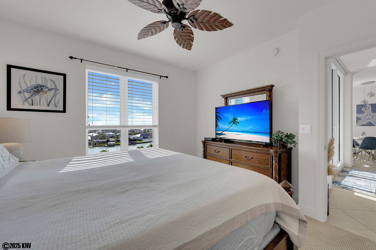Een slaapkamer met een groot bed, een plafondventilator, een televisie op een houten dressoir en een raam met zonwering.