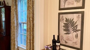 A room with a window, curtains, a framed botanical print, a small table with two bottles, and a trash bin.