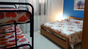 Un dormitorio con literas, una cama de madera, un ventanal con cortinas traslúcidas y una pared con un cuadro enmarcado.
