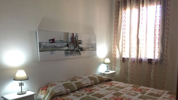 Un dormitorio con una cama, dos lámparas de noche, una mesita de noche, un cuadro en la pared y una ventana con cortinas.