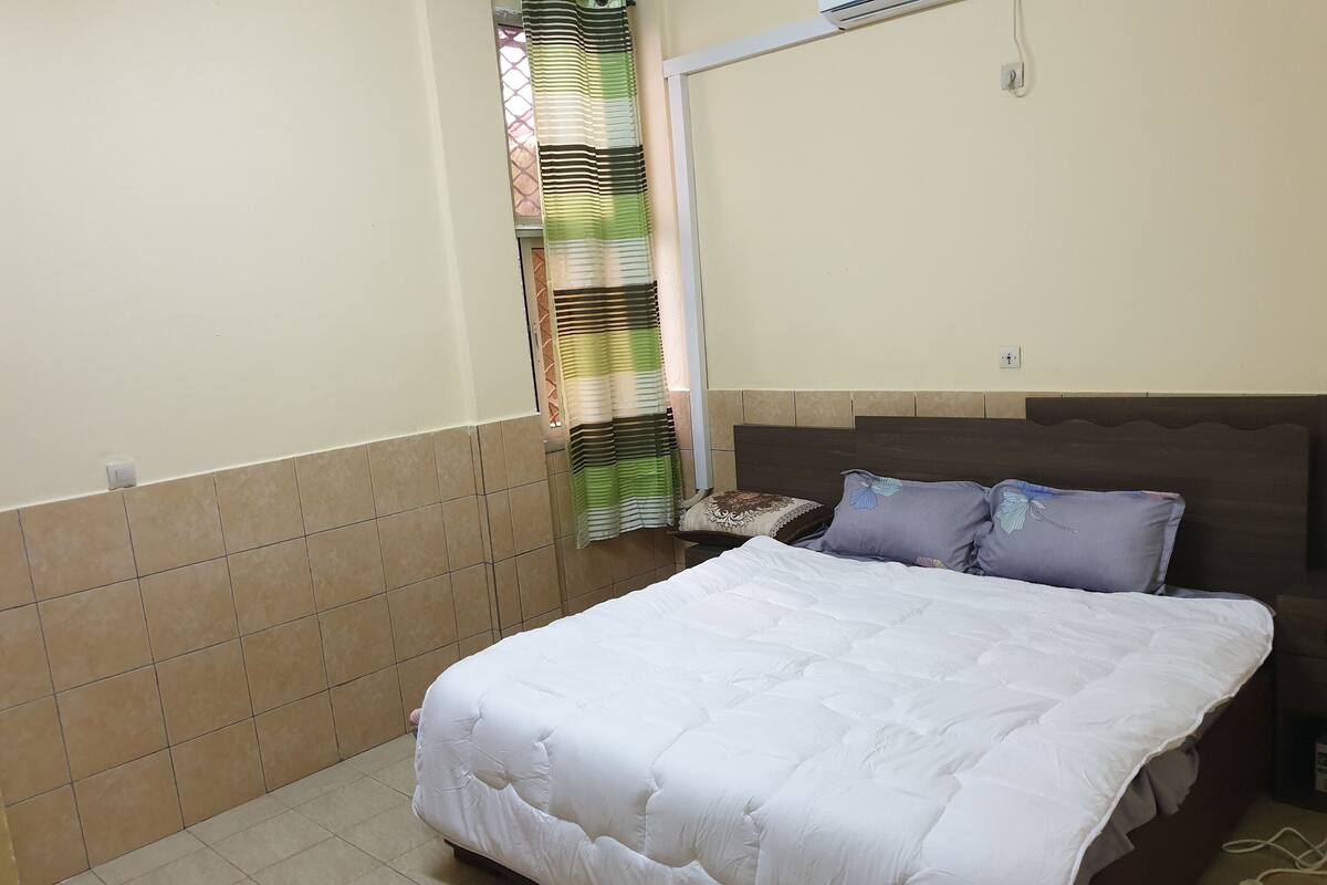 A neatly made bed with a white duvet and two pillows, a window with striped curtains, and a tiled floor.