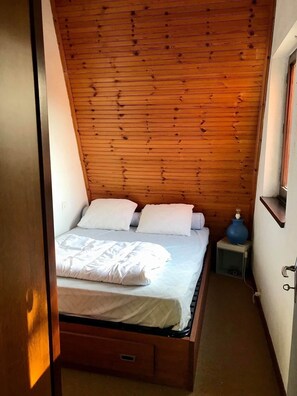 Ein kleines, gemütliches Schlafzimmer mit einem Holz-Kopfteil, einem Einzelbett mit weißer Bettwäsche, einem Nachttisch mit einer blauen Vase und einer kleinen Schublade unter dem Bett.