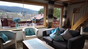 Un salon chaleureux avec vue sur un paysage montagneux, une table basse en bois, deux fauteuils et un canapé agrémenté de plusieurs coussins.