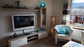 Un salon moderne doté d’un téléviseur à écran plat, d’un meuble de rangement en bois et d’un fauteuil confortable placé près d’une fenêtre offrant une vue sur l’extérieur.