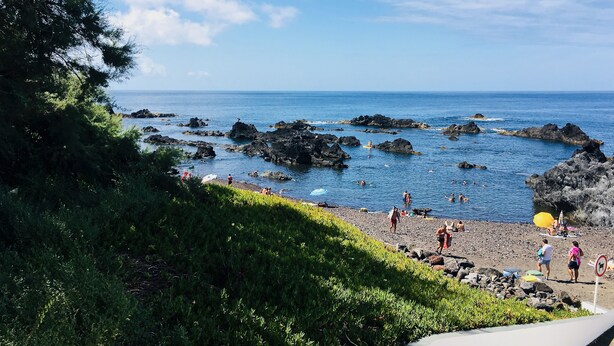 Une plage côtière avec un terrain rocheux, des gens qui se baignent et prennent des bains de soleil, et un ciel bleu dégagé.