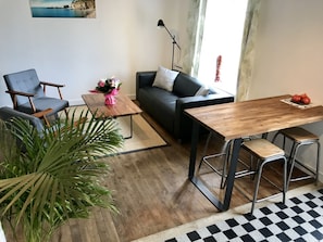A living room with a sofa, a chair, a wooden coffee table, and a dining table with stools.