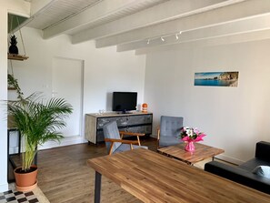 A living room with a wooden table, two chairs, a sofa, a TV on a wooden cabinet, a potted plant, and a framed picture on the wall.