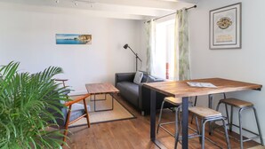 En kompakt opholdsstue med et træspisebord, to barstole, en sofa, en gulvlampe og en stor potteplante.