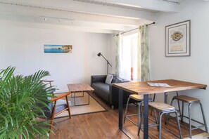 Un espacio habitable pequeño con una mesa de comedor de madera, dos taburetes tipo bar, un sofá, una lámpara de pie y una planta grande en maceta.