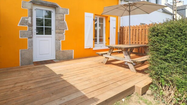 En træterrasse med et picnicbord og parasol, ved siden af en bygning med en lys orange facade og hvide skodder.