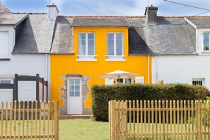 Una casa de dos pisos con una fachada amarillo brillante, una puerta blanca y un pequeño jardín con cerca de madera.