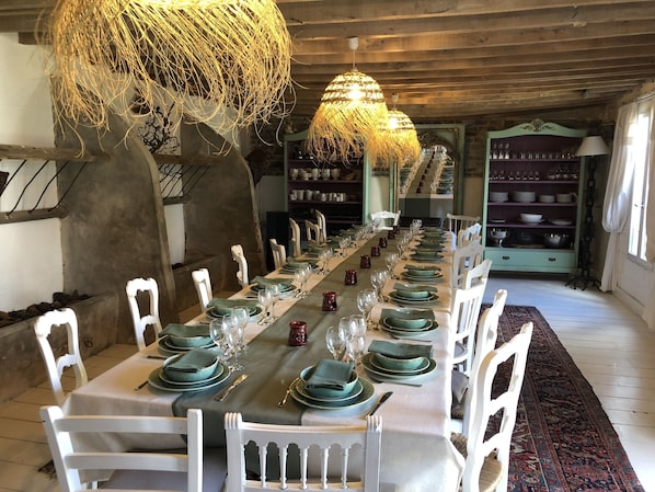 Mesa de jantar comprida posta com pratos verdes, cadeiras brancas e luminárias penduradas.