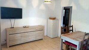 A small, well-lit room with a wooden cabinet, a white refrigerator, a dining table with chairs, and a flat-screen TV mounted on the wall.