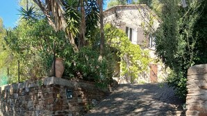 A narrow, cobblestone street flanked by stone walls and lush greenery, leading to a building with a tiled roof.