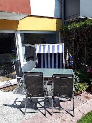 Een terras buiten met een glazen tafel en twee stoelen, een gestreept zonnescherm en een kleine tuin.