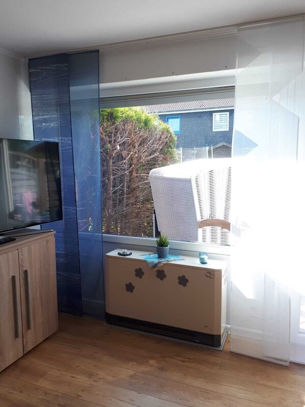 Ein Zimmer mit Fernseher auf einem Holzschrank, einem Fenster mit Jalousien, einem Korbsessel und einem kleinen Tisch mit Pflanze.