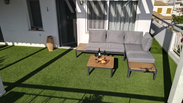 A modern outdoor seating area with a grey sofa and wooden tables, surrounded by artificial grass.