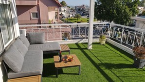 Een balkon met een grijze bank, een houten tafel met een fles en fruit, en uitzicht op een stadsgezicht.