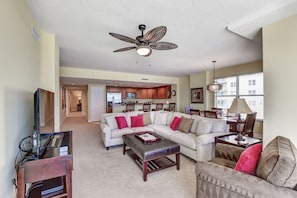 A spacious living room with a sectional sofa, a coffee table, and a TV on a wooden stand. A ceiling fan is visible above.