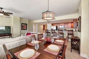 A dining area with a wooden table set for six, a ceiling fan, and a kitchen in the background.