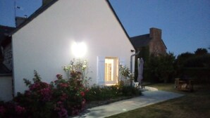 A white building with a lit entrance, surrounded by a garden with pink flowers. - The charm of the Breton countryside (Quemper Guezennec)
